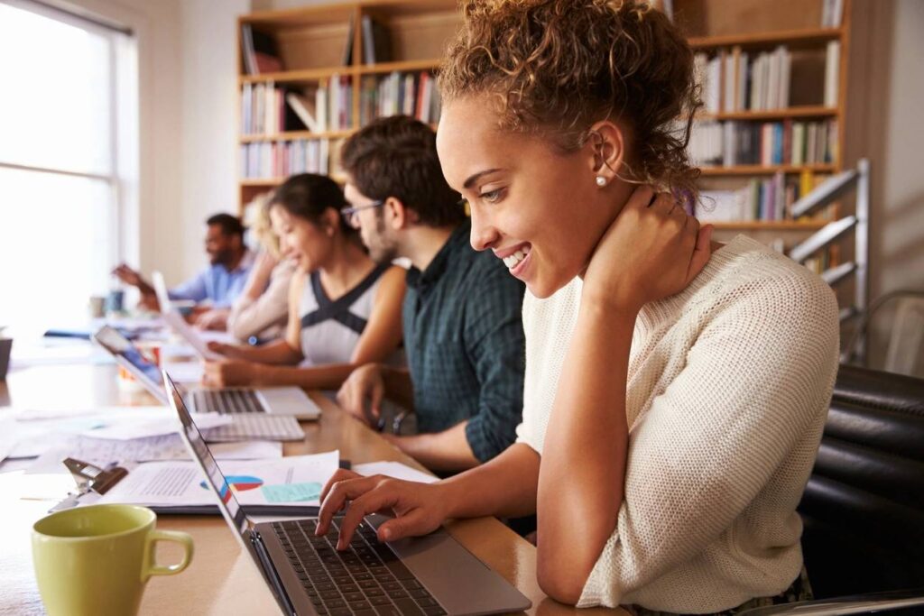 Jobseekers immersed in a focused atmosphere, sitting at a desk and using laptops. A smiling girl in the foreground looks at her laptop,looking for job oportunities in the UK, conveying a welcoming and supportive environment for job search and CV uploads