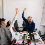 Four individuals celebrating success in a professional setting. Arms raised in joy, the business environment includes a table with laptops and various office essentials, embodying the core principles of strategic staffing solutions for enhanced workplace performance.