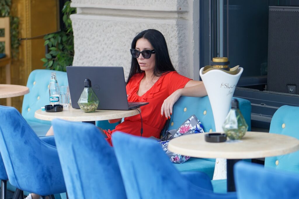 A woman sitting in a cafe with sunglasses, using a laptop. She appears relaxed and focused, representing the concept of remote work while enjoying a coffee shop ambiance.